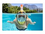 Woman snorkeling in a pool with a full-face mask, mountains and trees in background.