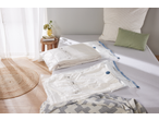 Two vacuum storage bags with textiles on a bed, next to a stool with a vase and books.