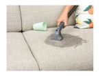 Hand cleaning a sofa with a spot cleaner, next to a green cup and a floral cushion.