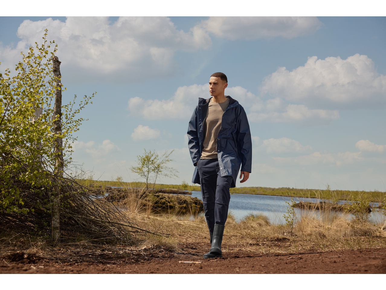 Man in een marineblauwe trenchcoat en donkere broek wandelt buiten bij een meer.