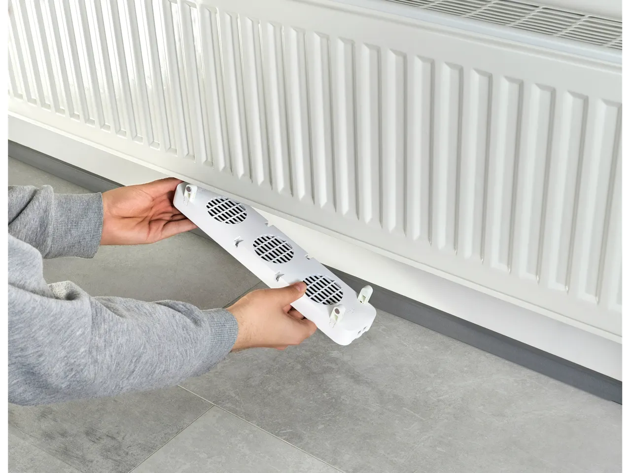 Hands holding a white SILVERCREST® Radiator Booster with three fans, positioned near a white radiator.