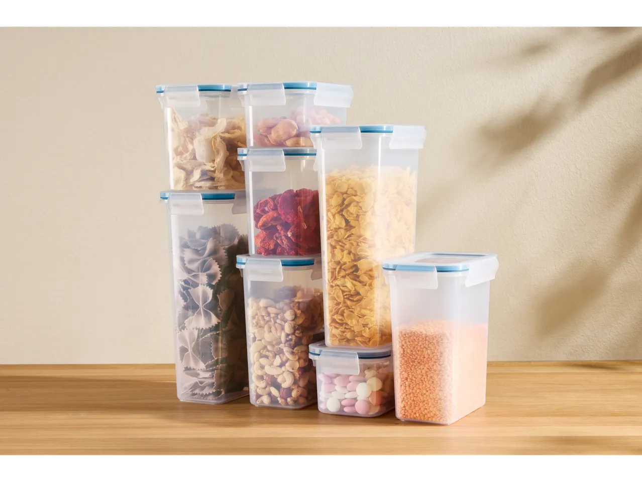 Διαφανή δοχεία αποθήκευσης τροφίμων γεμάτα με ζυμαρικά, αποξηραμένα φρούτα, ξηρούς καρπούς, δημητριακά και φακές σε ξύλινη επιφάνεια.