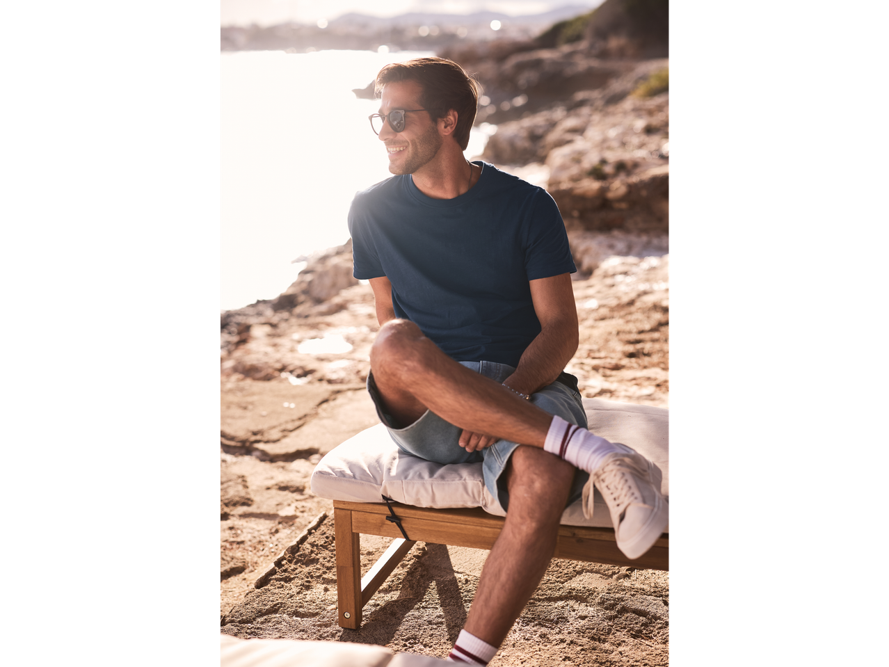 Bărbat relaxat pe o șezlong lângă mare, îmbrăcat cu un tricou bleumarin și pantaloni scurți.