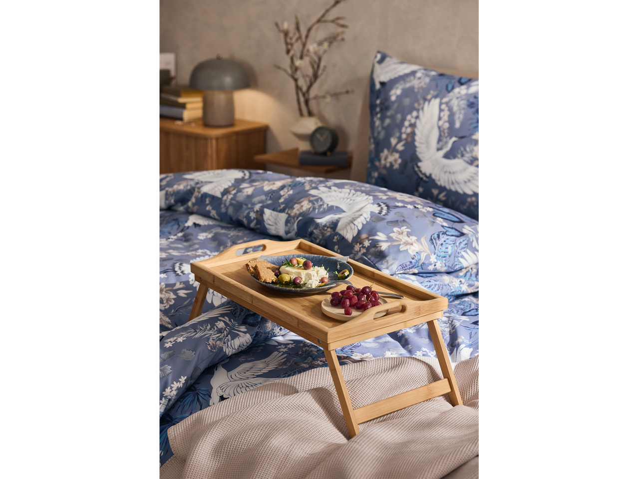 A bamboo breakfast tray holds a plate of food and grapes on a bed with blue and white bedding.