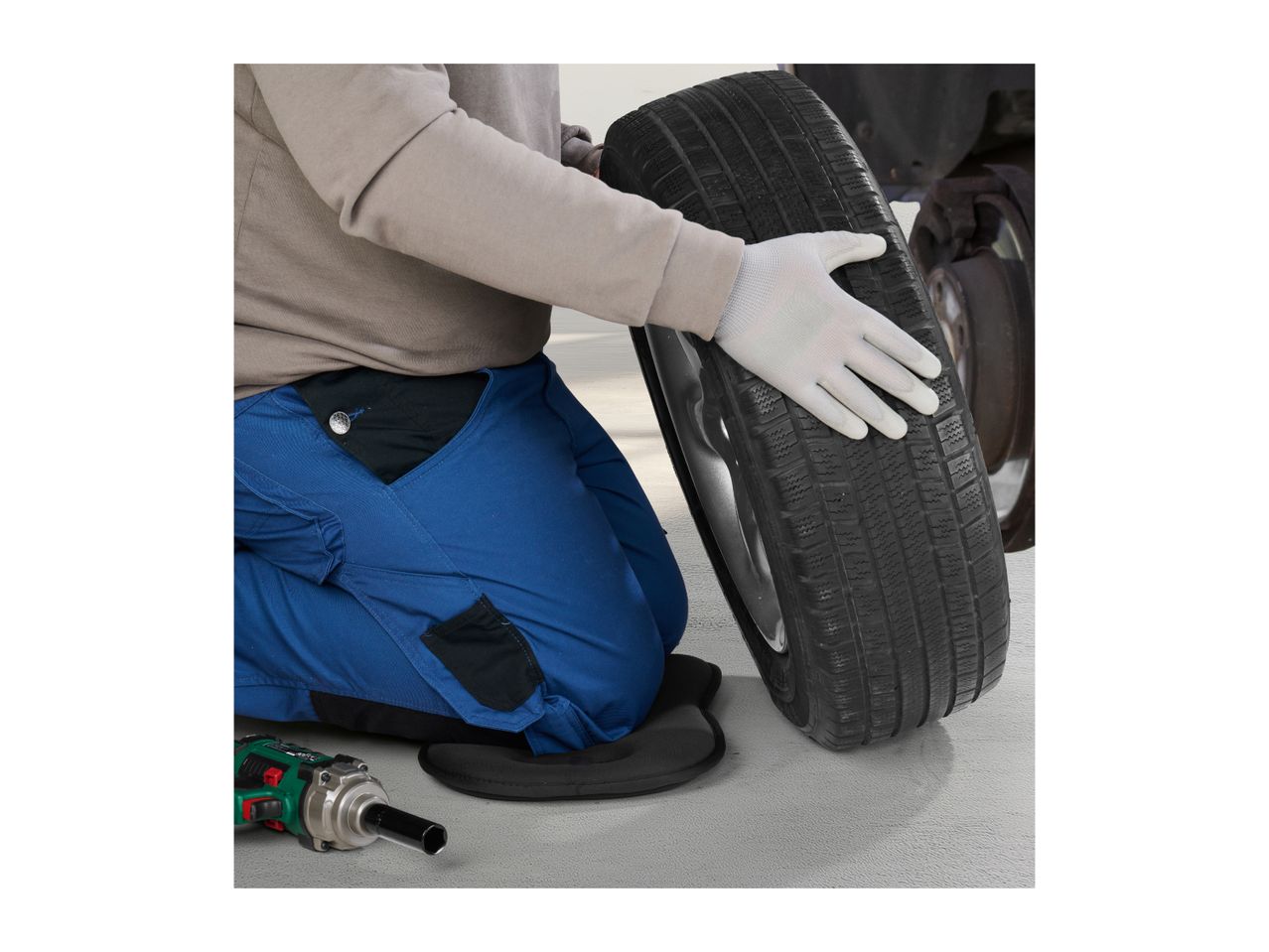 Mechanic kneeling on a knee pad, wearing work gloves, holding a car tire, with an impact wrench nearby.