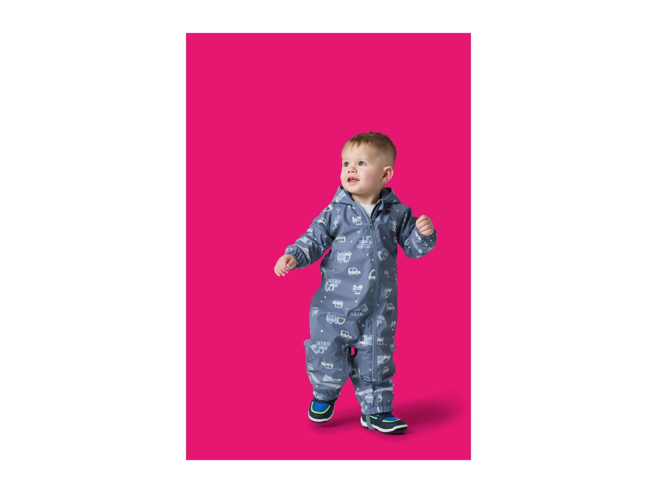 Toddler in a blue rain suit with vehicle patterns, standing on a pink background.