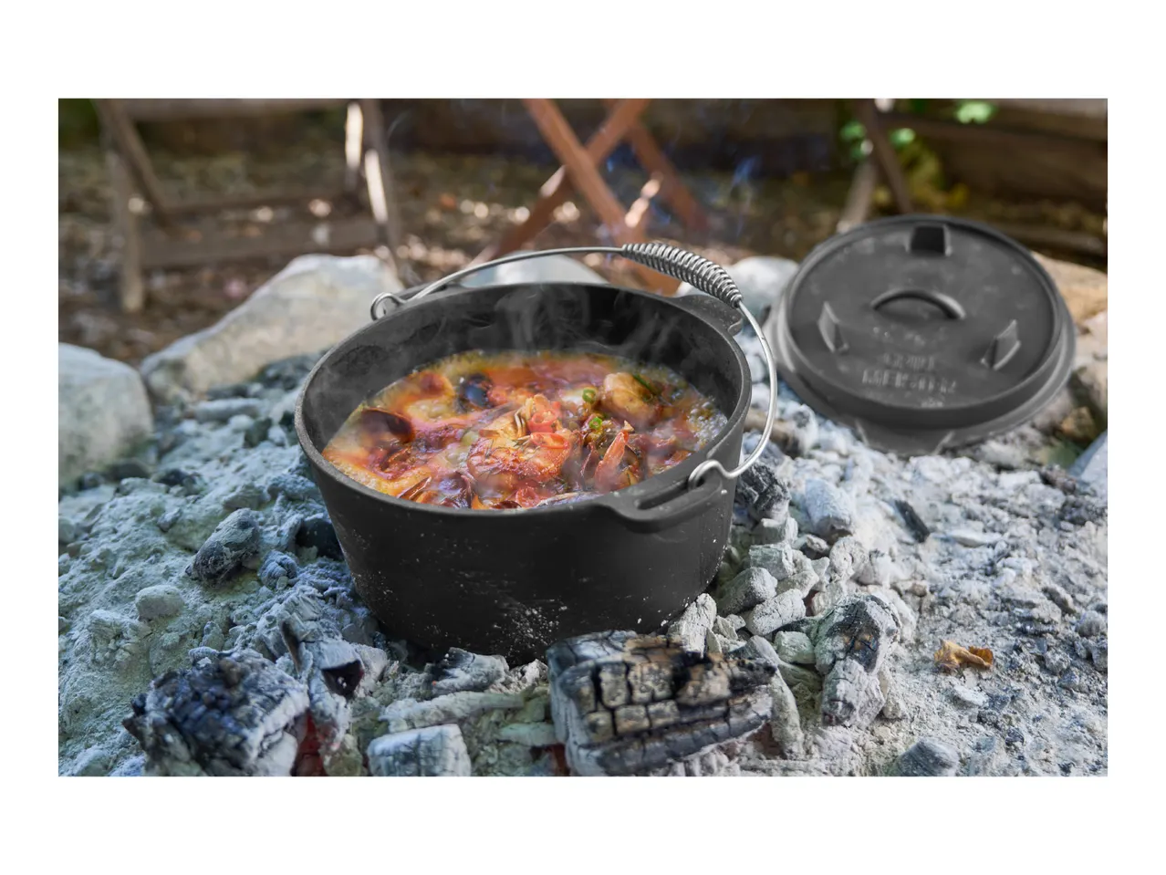 Dutch oven with seafood stew cooking over hot coals outdoors.