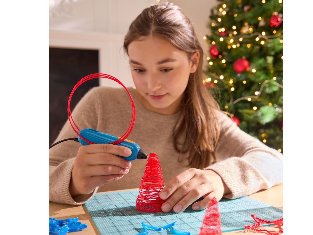 Момиче използва 3D писалка, за да създаде червена коледна елха, с коледно дърво на заден план.