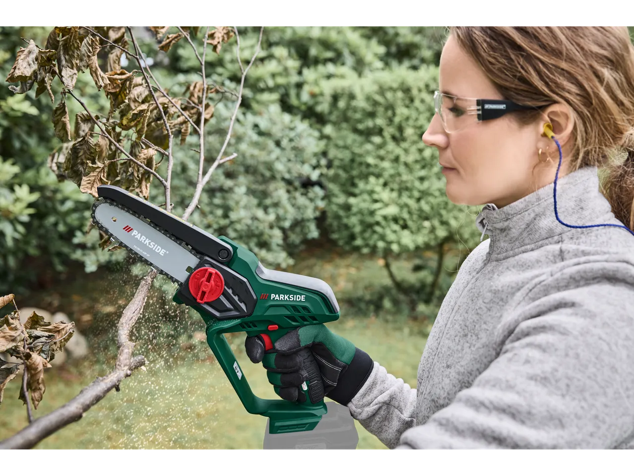 Woman cutting a branch with a cordless wood cutter, wearing safety glasses and earplugs.