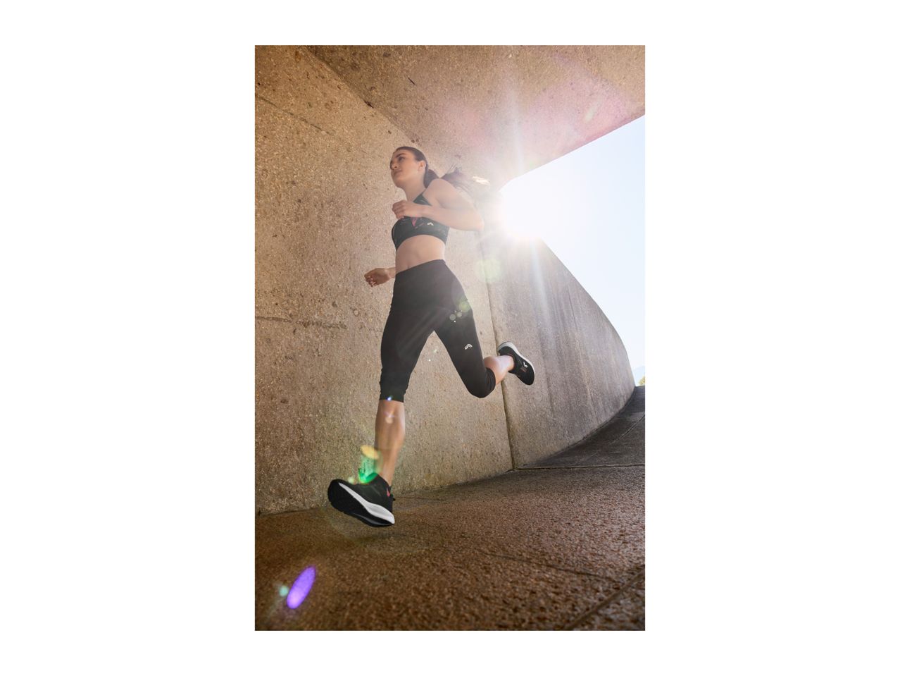 Woman running in a black sports bra and black capri leggings with sun flare.