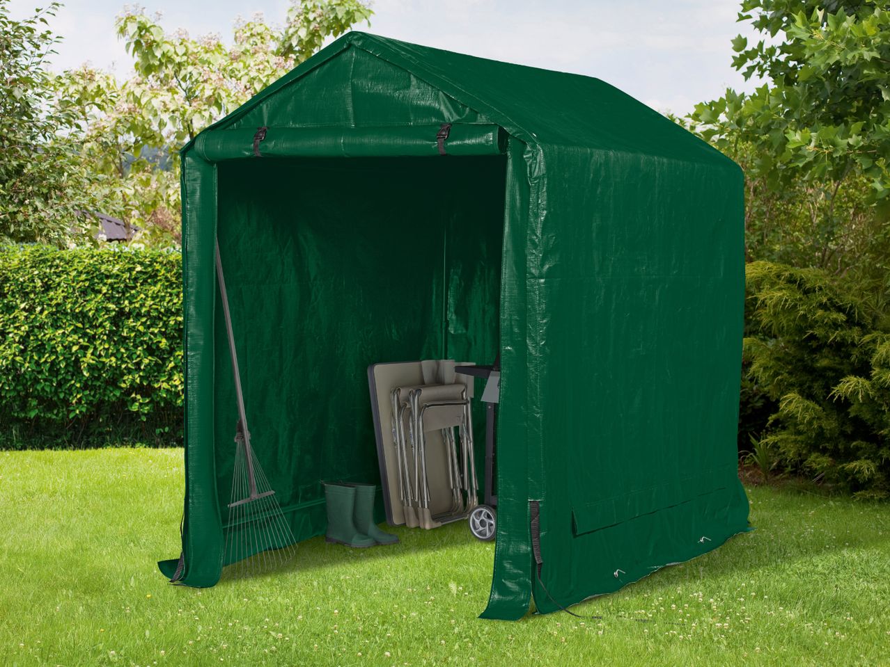 Green garden storage tent with a rake, boots, and folded chairs inside on a lawn.