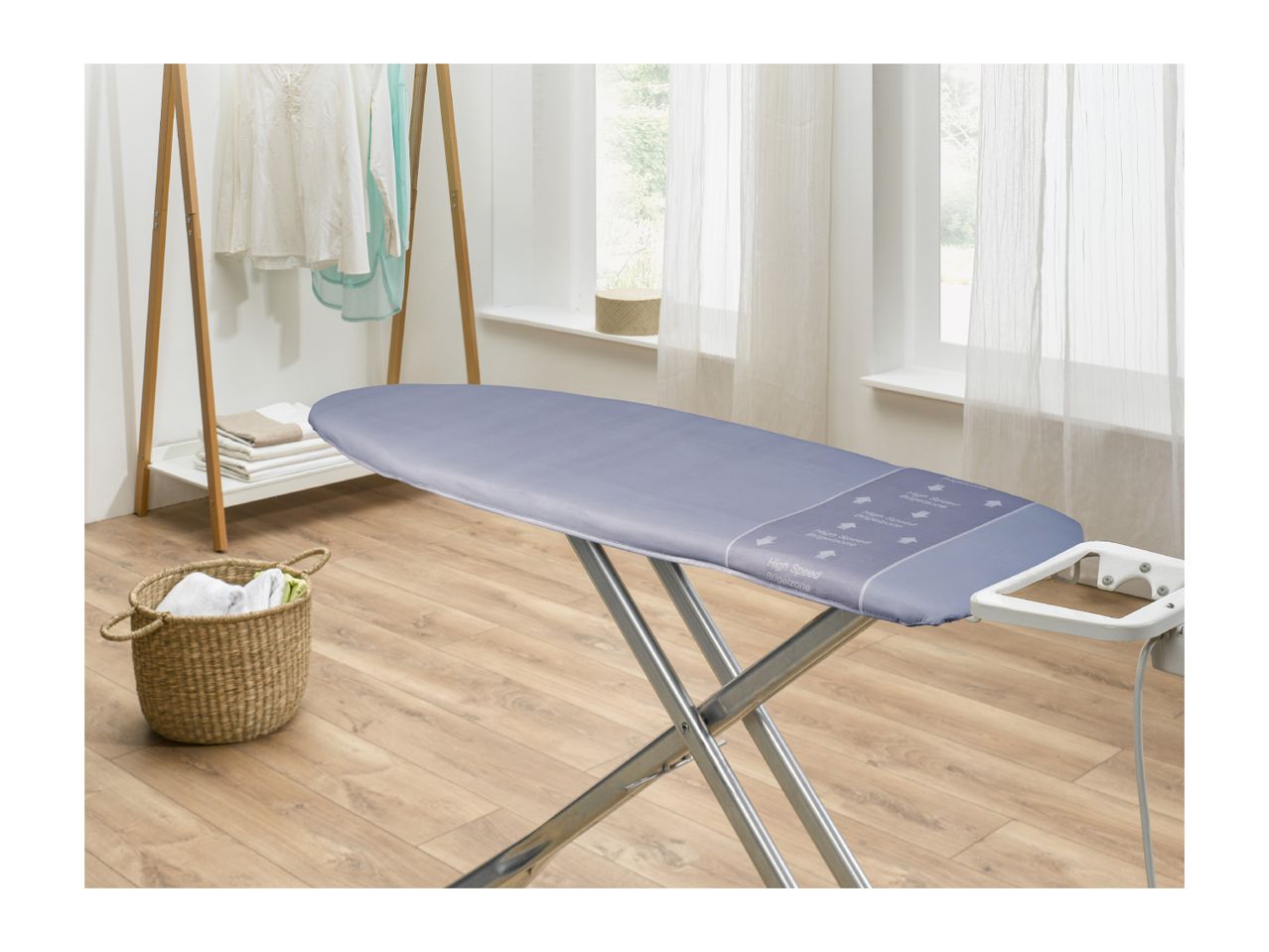 Ironing board with a blue cover, laundry basket, and clothes rack in a bright room.