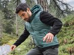 Man in a teal puffer vest and black long-sleeved shirt hiking in a forest.
