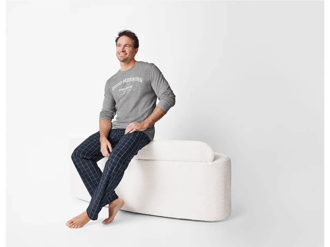 Lachende man in grijze en blauwe pyjama, zittend op een witte bank.