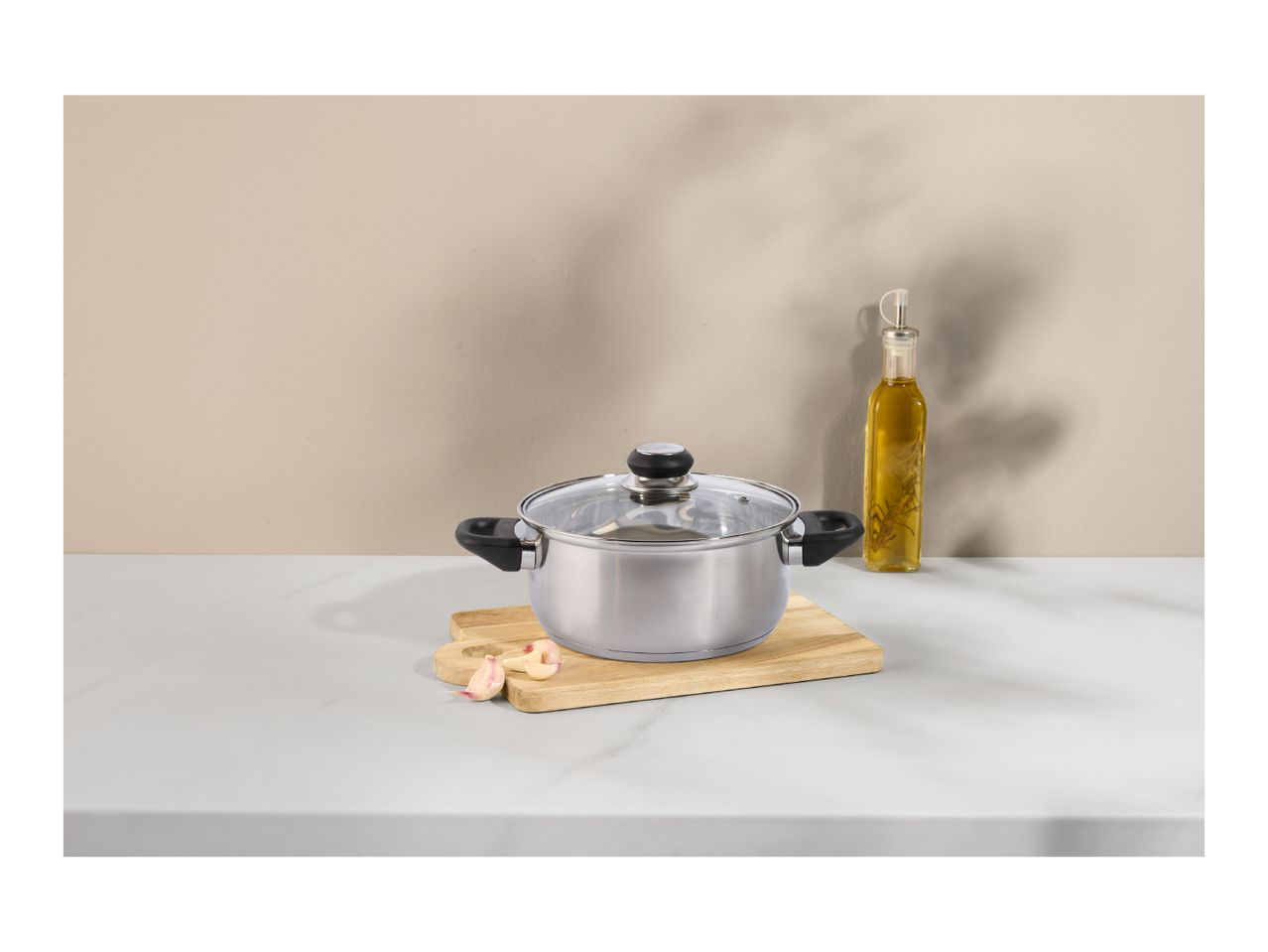 Stainless steel pot with glass lid on a wooden board, with garlic and olive oil.