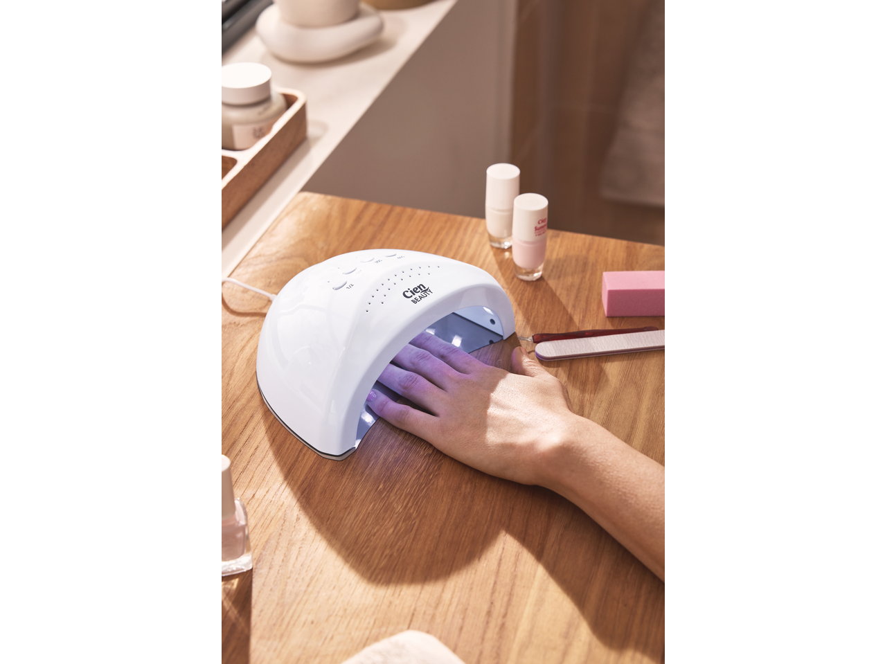 A hand under a nail lamp, with nail polish bottles and manicure tools on a wooden table.