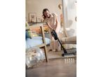 A woman uses a black cordless vacuum cleaner on a light wood floor.
