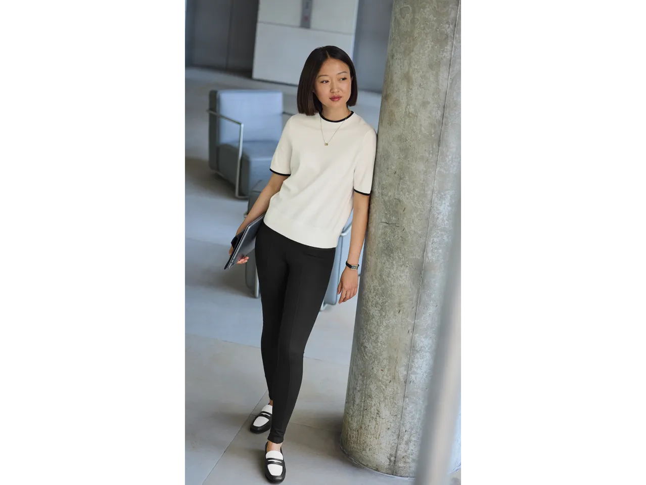 Mulher elegante com suéter branco e calça preta, com laptop e sapatos bicolor.