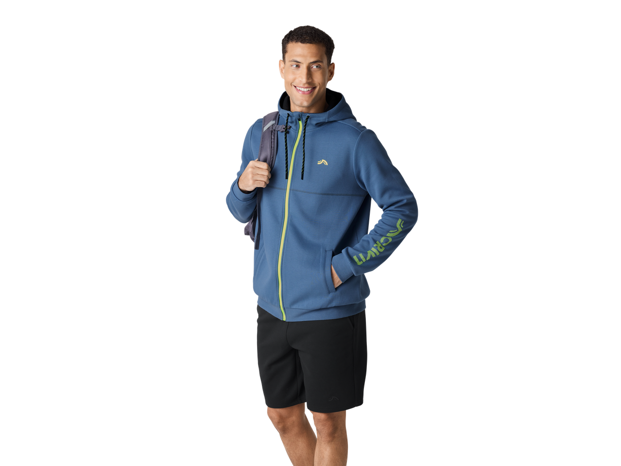 Smiling man in a blue CRIVIT zip-up jacket and black shorts, carrying a backpack.