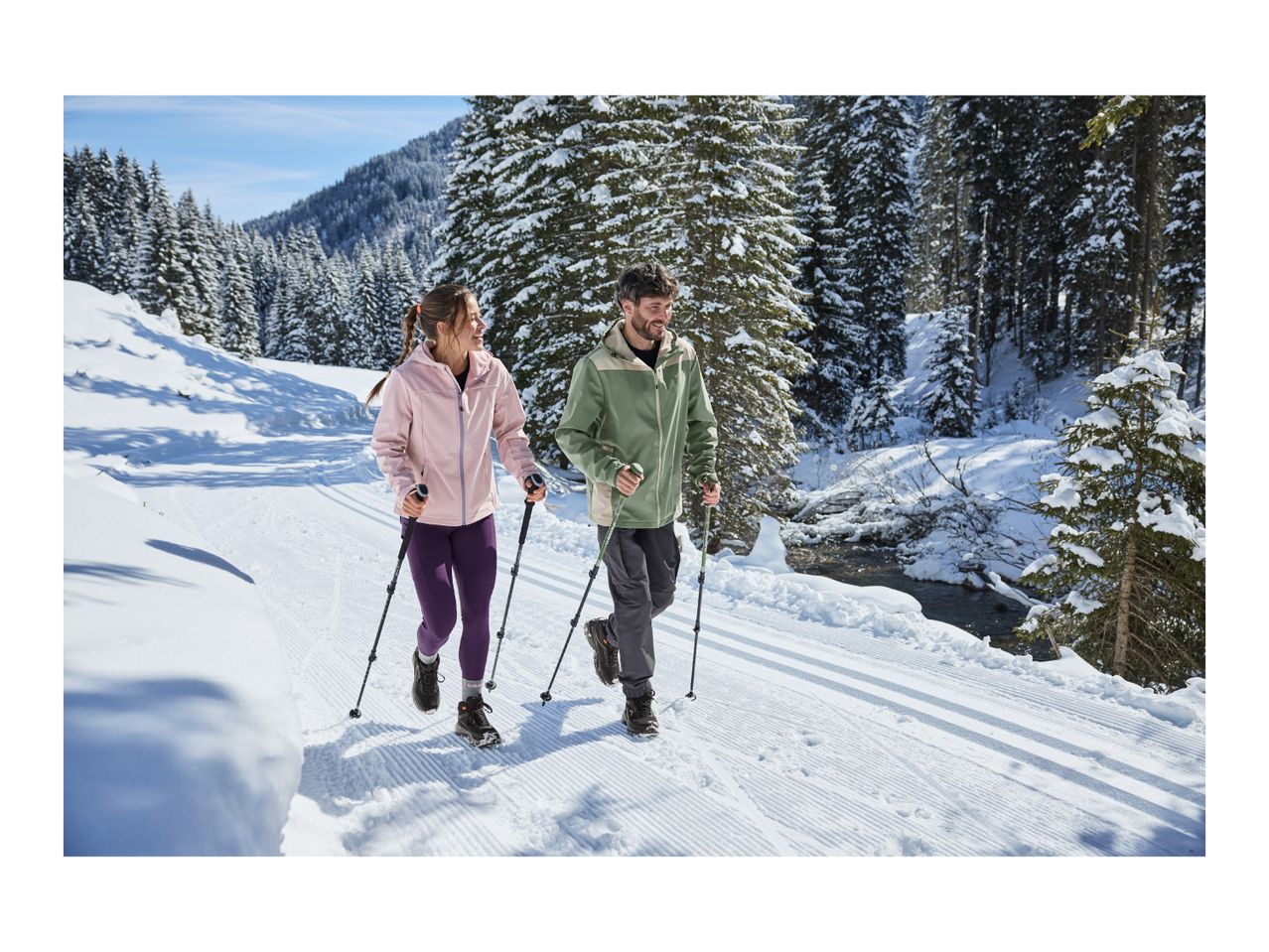 A man and a woman winter hiking with trekking poles on a snowy trail.