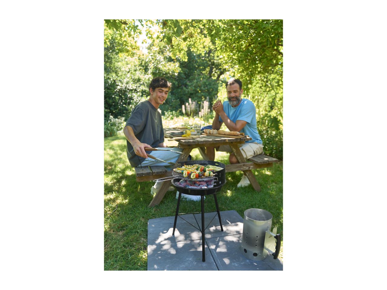 Two men grilling vegetable skewers on a barbecue in a garden.