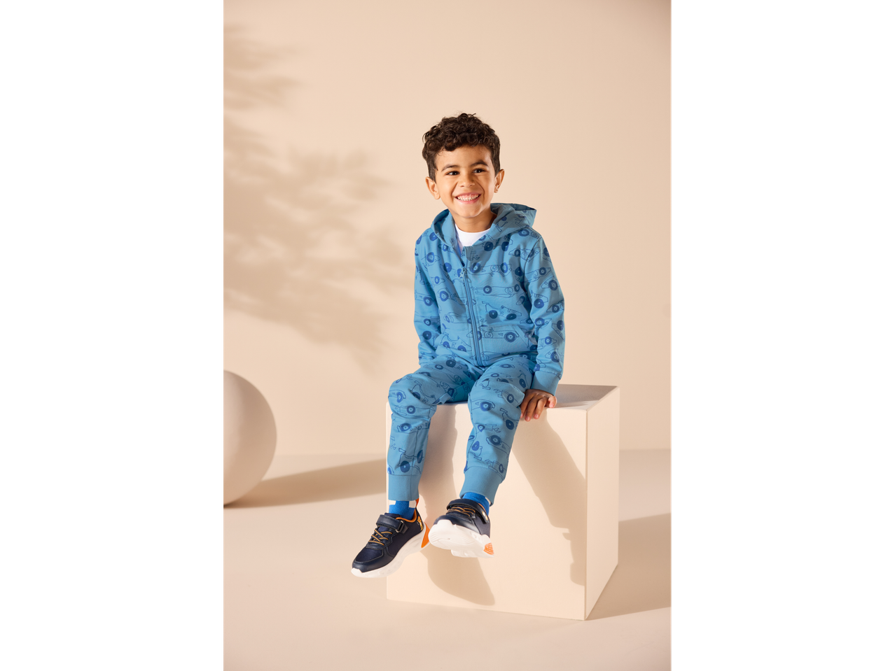 Nasmiješeni dječak u plavoj Lupilu® trenirci s printom automobila i tamnoplavim tenisicama.
