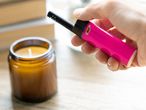 A hand holds a pink long lighter to light a candle in an amber jar.