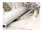 Person vacuuming under a light grey sofa with a cordless stick vacuum cleaner on a rug.