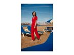 Woman in red jumpsuit walking barefoot on a sandy beach with blue lounge chairs.