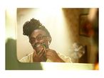 Man with dreadlocks and glasses shaving with an electric shaver in a mirror.