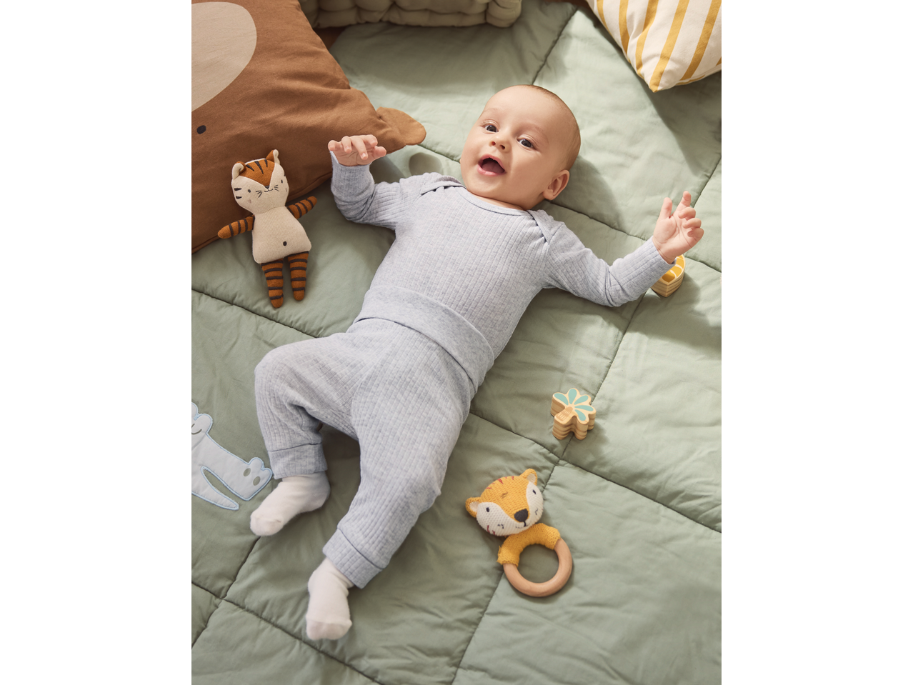 Baby in light blue ribbed sleepsuit, lying on a green blanket with soft toys and wooden rattles.