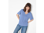 Woman in a light blue V-neck t-shirt and blue jeans, leaning against a white wall.