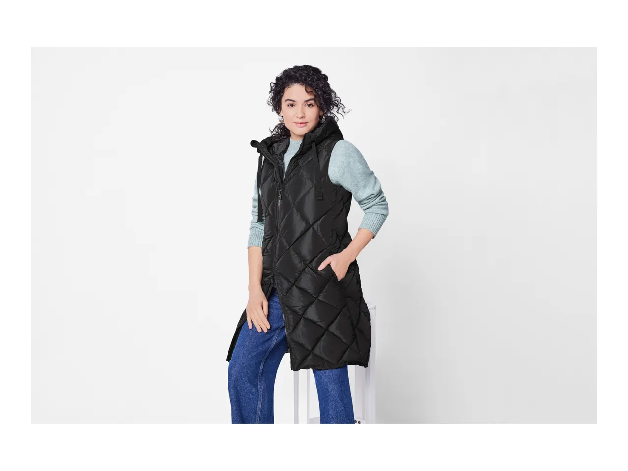 Woman in a black quilted longline vest, light blue sweater, and blue jeans.