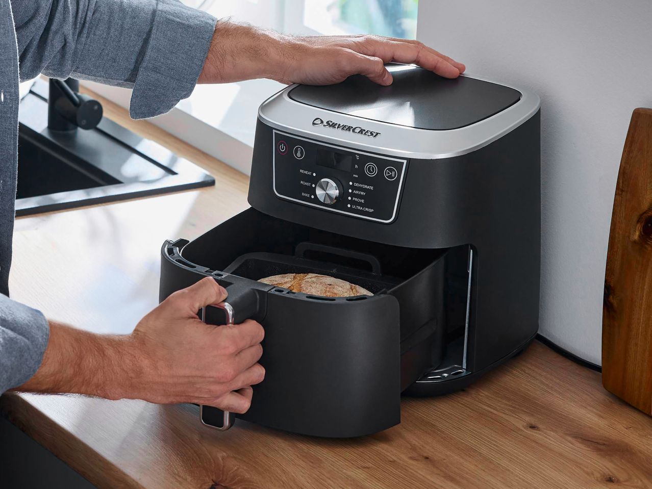 A person operating a black air fryer with a digital display showing cooking modes like REHEAT, ROAST, BAKE, DEHYDRATE, AIR FRY, PROVE, and ULTRA CRISP.
