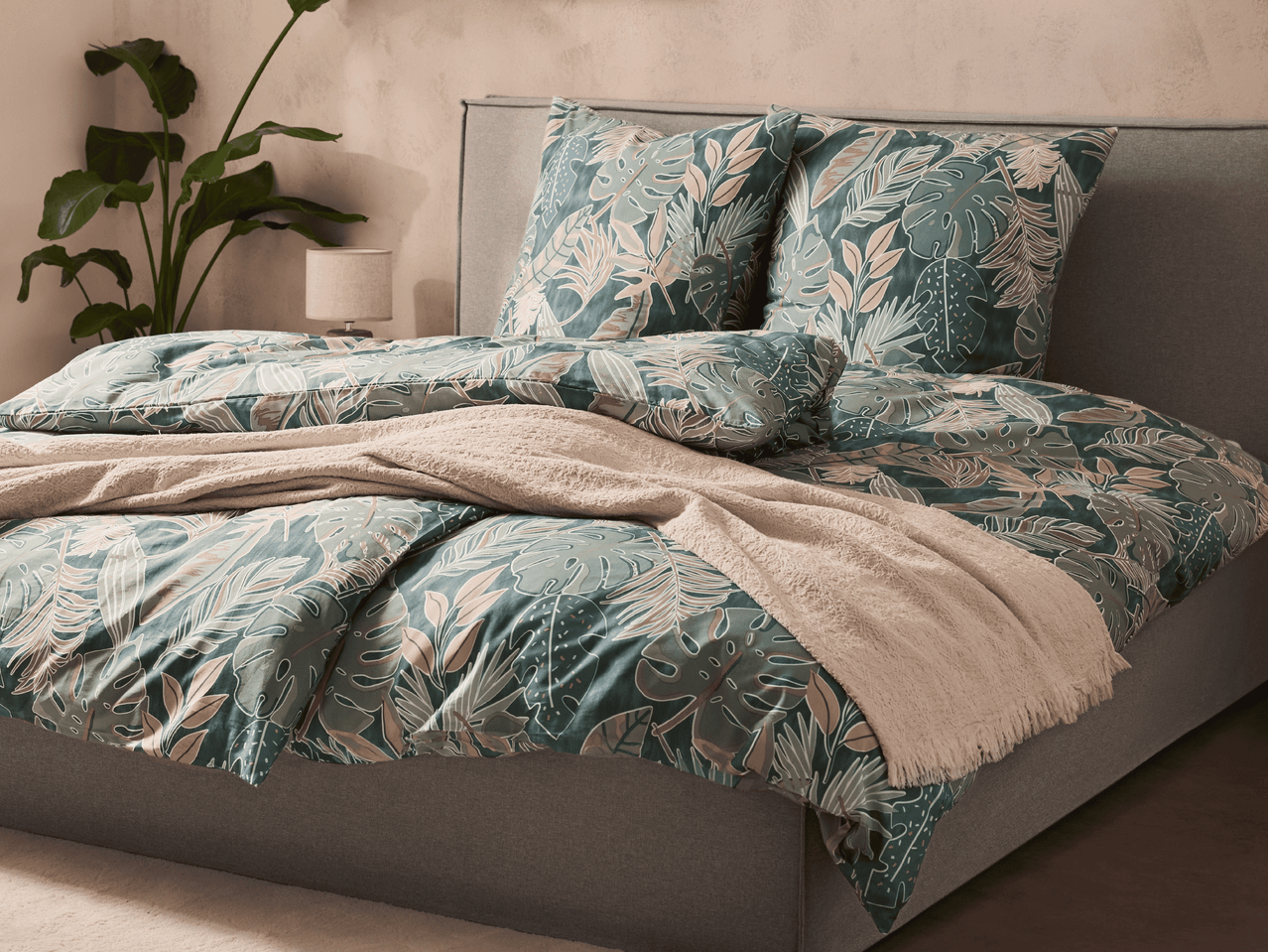 Bett mit Bettwäsche im tropischen Blättermuster, einem beigen Überwurf und einer Pflanze.
