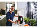 Homem sorrindo enquanto coloca batatas-doces fritas em uma fritadeira de ar na cozinha moderna.