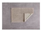 A beige bath mat with a textured surface, partially folded to show its underside, on a gray tiled floor.