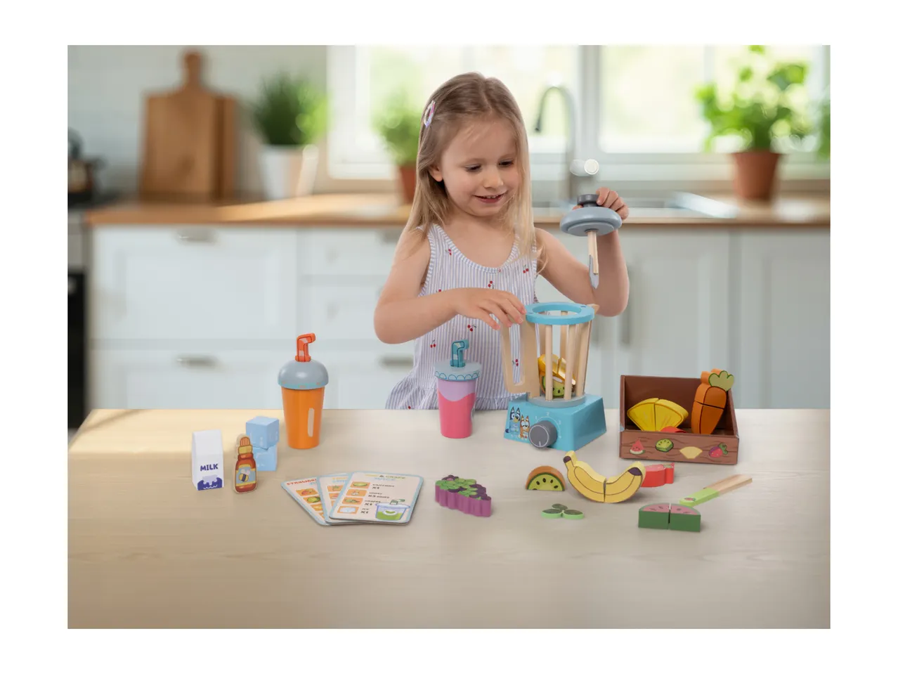 A young girl playing with a wooden toy blender set, including fruit, cups, and recipe cards.