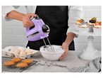A person uses a purple hand mixer to whip egg whites in a glass bowl, surrounded by baking ingredients and cupcakes.