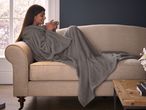 Woman relaxing on a sofa with a grey wearable blanket, holding a mug.