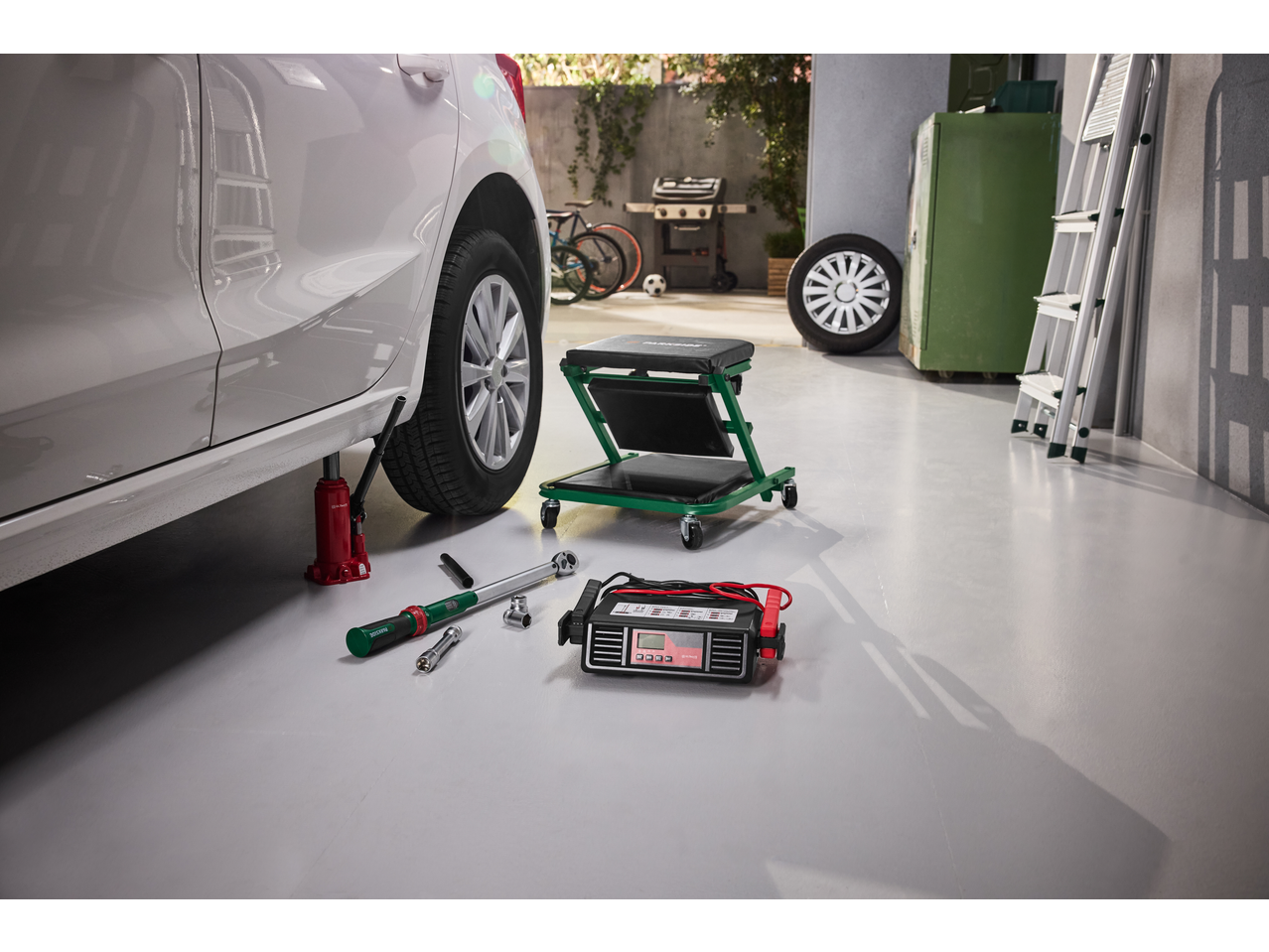 Car jacked up for tire change, with a torque wrench, battery charger, and mechanic's creeper on the garage floor.