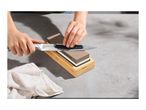 Hands sharpening a knife on a two-sided whetstone with a wooden base.