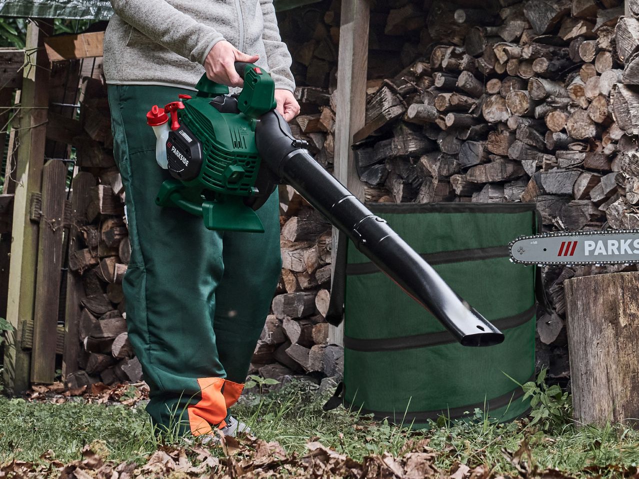 Person holding a leaf blower, with a chainsaw and stacked firewood in the background.
