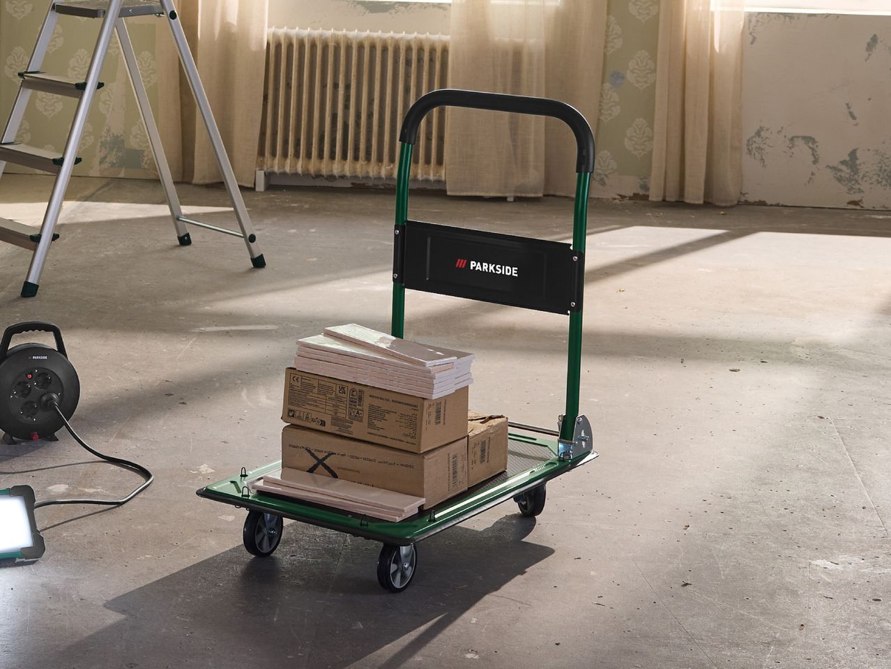 Hand truck with boxes of tiles and a power reel in a room under renovation.