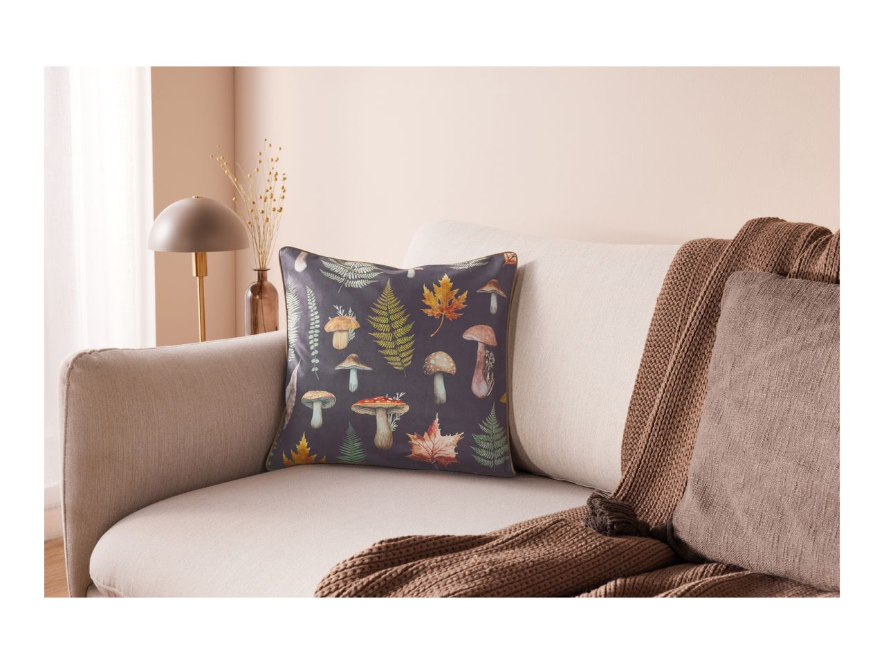 A mushroom and fern patterned pillow on a sofa with a brown knitted throw.
