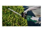 Person trimming a green hedge with a cordless hedge trimmer, wearing protective gloves.