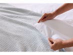 A person's hands are shown making a bed with a light blue textured throw blanket and white bedding.