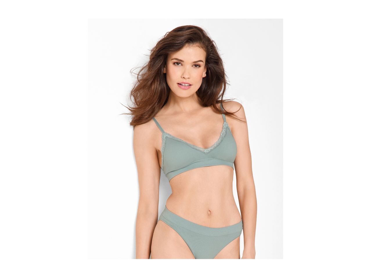 Woman in a light green bralette and matching briefs, leaning against a white wall.