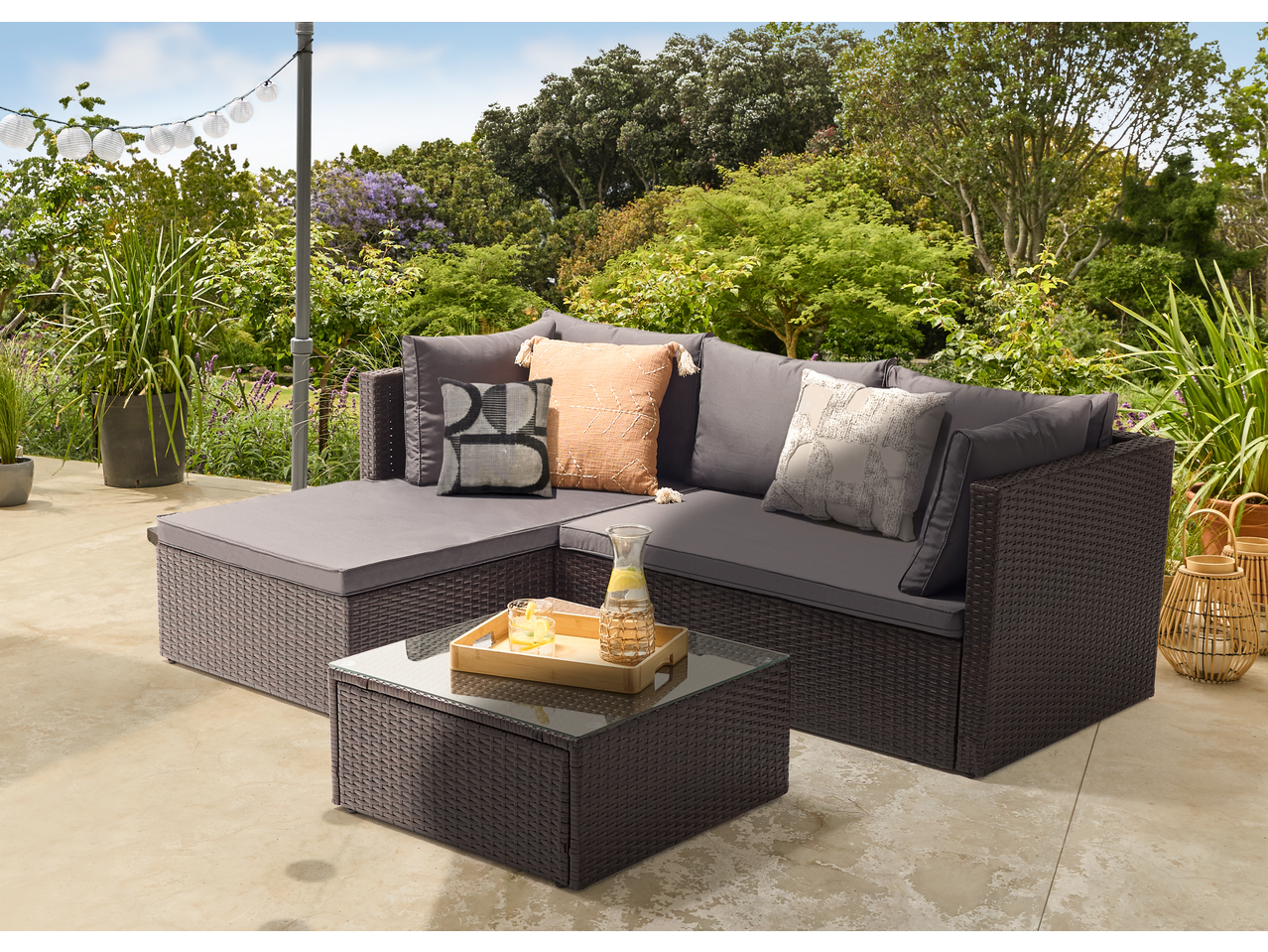 Outdoor rattan corner sofa with grey cushions and a glass-top coffee table on a patio.
