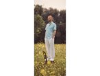 A man in a light blue linen shirt and white pants stands on a chair in a field of yellow flowers.